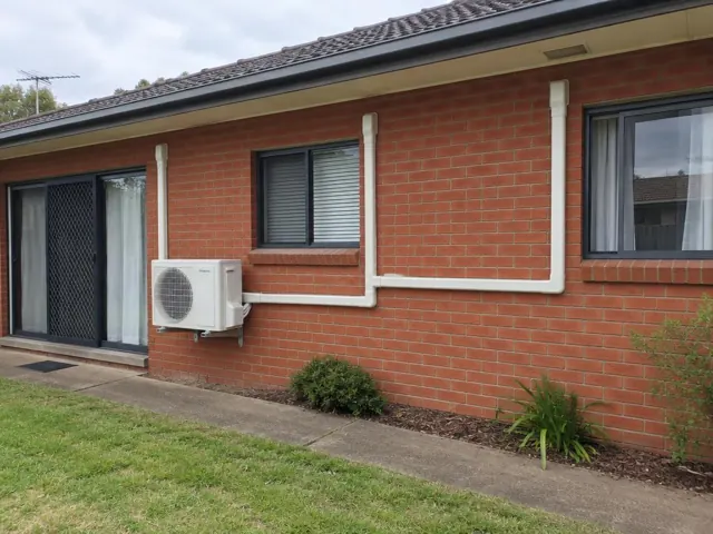 Brick house wall with a wall-mounted outdoor air conditioner unit and white pipe covers in Pooraka